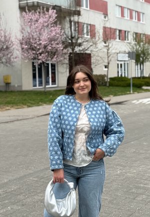 Joven mujer con chaqueta azul de lunares y vaqueros, sosteniendo un bolso plateado, de pie en la acera cerca de árboles en flor y edificios residenciales.