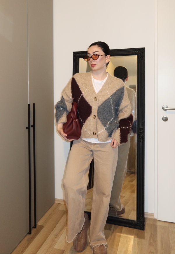 Vrouw draagt een oversized beige, grijze en bordeauxrode cardigan, beige broek, bruine schoenen, zonnebril, houdt een bordeauxrode handtas vast en staat voor een spiegel.