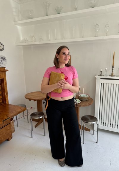 Joven mujer con camisa rosa y pantalones negros sosteniendo un bolso mostaza, de pie en una habitación blanca con muebles de madera y metal y cristalería en las estanterías.