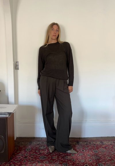 Suéter negro de manga larga con corte holgado, combinado con pantalones de pierna ancha negros de cintura alta y zapatos metálicos grises sobre una alfombra de patrón.