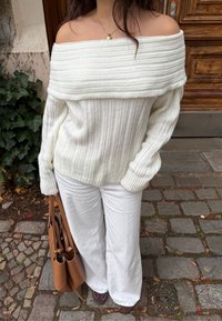 Fehér bordázott, váll nélküli pulóver hosszú ujjakkal, fehér bő szárú nadrággal és barna kézitáskával párosítva. Természetes kő hátterű.