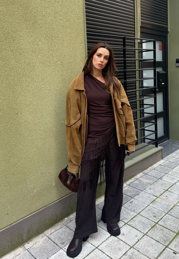 Žena v hnědém volném kabátu, tmavých střapatých šatech, černých širokých kalhotách a botách stojí na chodníku vedle zelené zdi a černých kovových vrat.