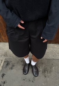 Personne portant un short noir, des chaussures noires, des chaussettes blanches et un haut à manches longues, debout sur une surface en béton près d'une porte en bois.