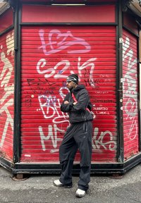 Homme portant une veste noire, un pantalon, un bonnet et des lunettes de soleil, debout les bras croisés devant un volet métallique de magasin rouge couvert de graffiti.