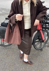 Braune Wildlederjacke, weißes geripptes Shirt, braune Lederrock, braune Umhängetasche und braune Schuhe mit weißen Socken, neben einem Fahrrad.