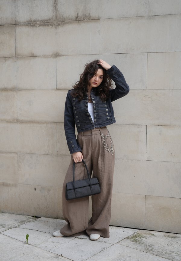 Pitkäkiharahiuksinen nainen pukeutuneena lyhytmusta takkiin, leveät beigeihin housuihin, joissa on ketju, musta käsilaukku kädessään, seisoo kiviseinää vasten.
