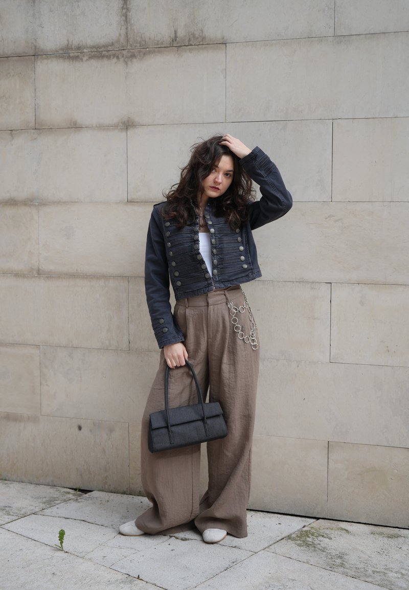 Γυναίκα με μακριά κατσαρά μαλλιά, που φοράει μαύρο κοντό μπουφάν, φαρδιά μπεζ παντελόνια με αλυσίδα, κρατώντας μια μαύρη τσάντα, στέκεται σε έναν λιθότοιχο.