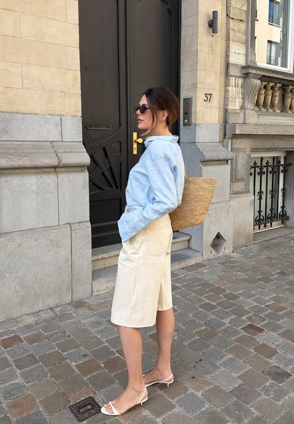 Femme portant une chemise bleu clair, un short crème, des sandales blanches et des lunettes de soleil, se tient dans une rue pavée près d’une porte noire et d’un bâtiment en pierre.