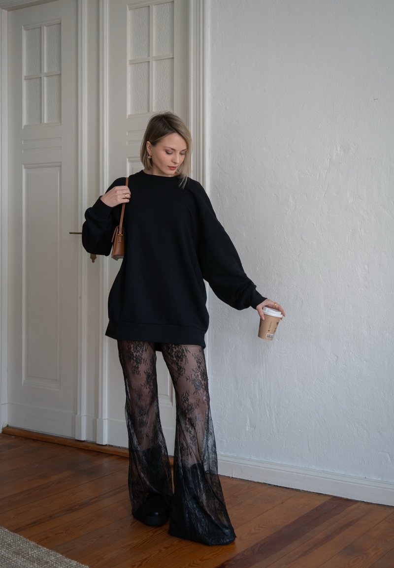 Μαύρο oversized φούτερ με φαρδιές φουσκωτές μανίκες, σε συνδυασμό με διάφανες μαύρες δαντελένιες παντελόνες σε φαρδιά γραμμή. Κρατώντας ένα φλυτζάνι καφέ, στέκεται σε ξύλινο πάτωμα.