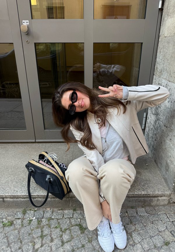 Jeune femme en tenue beige et lunettes de soleil assise sur une marche en pierre devant une porte en verre, faisant le signe de la paix de la main droite à côté d’un sac noir.
