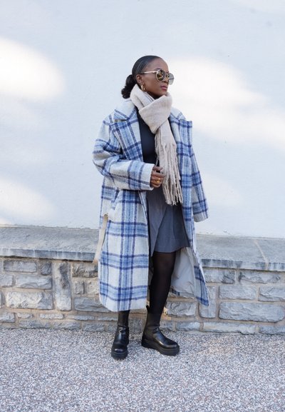 Abrigo de cuadros en gris claro y azul, diseño oversized, mezcla de lana; bufanda beige que cae, vestido gris, botas de plataforma negras, contra un muro de piedra.