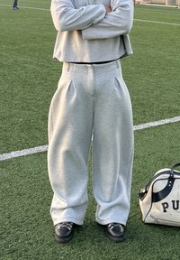 Pantalon de survêtement gris à jambes larges avec plis et surface texturée. Associé à des chaussures noires à bout arrondi, posé sur un gazon vert.