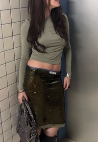 Olivenfarvet langærmet top kombineret med en skinnende grøn palietnederdel med blå taljebånd og et sølvfarvet armbånd. Mørk håndtaske i hånden.