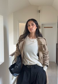 Trench-coat beige porté sur un pull crème, pantalon noir taille haute et sac à main en cuir noir. Des bijoux simples complètent le look.