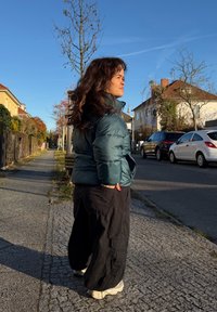 Femme aux cheveux bouclés portant une doudoune bleu canard et un pantalon cargo noir, debout sur un trottoir ensoleillé d'une banlieue, regardant vers la droite.