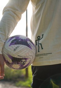 Λευκό πουκάμισο με μακριά μανίκια και πράσινο γραφικό σχέδιο στο πλάι, κρατώντας μια ποδοσφαιρική μπάλα με μωβ λεπτομέρειες και μαύρα-άσπρα σχέδια.