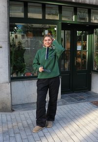 Jersey polar verde con cuello alto, botones oscuros y un bolsillo lateral, combinado con pantalones negros y calzado beige, de pie al aire libre.