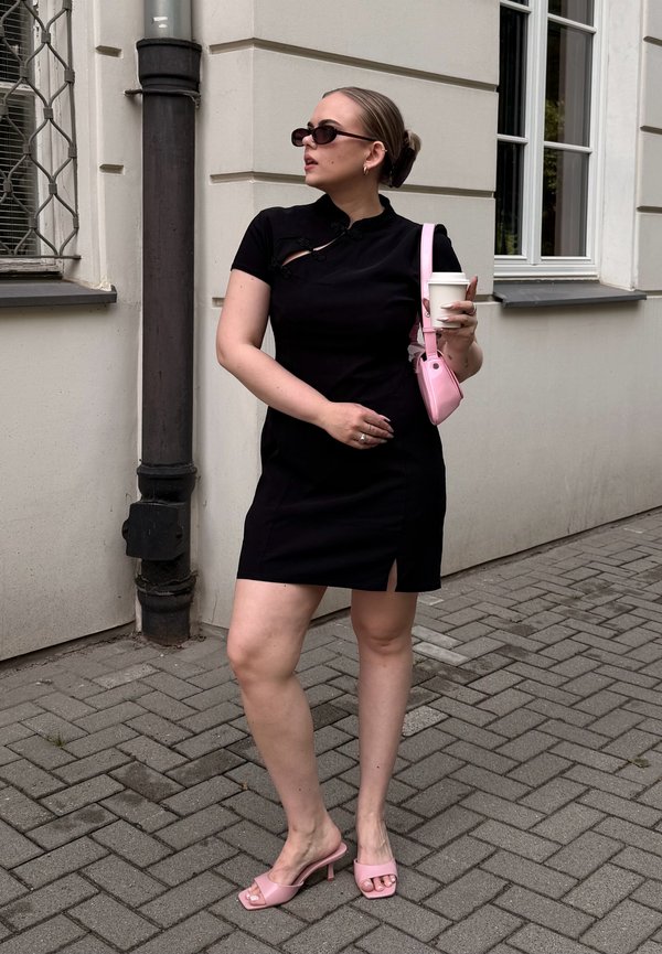 Mujer con vestido negro, llevando gafas de sol, sosteniendo una taza de café y un bolso rosa, de pie en una acera pavimentada junto a un edificio.