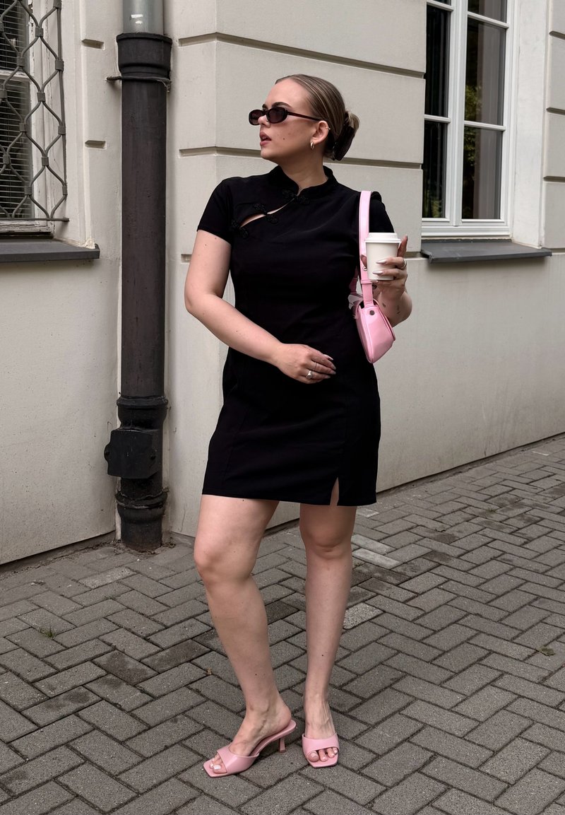 Femme en robe noire portant des lunettes de soleil, tenant une tasse de café et un sac rose, debout sur un trottoir pavé près d'un bâtiment.