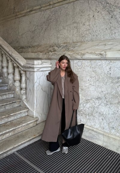 Abrigo oversized marrón, suéter de punto beige, pantalones negros, zapatillas grises y un bolso tote negro. Fondo de escalera y pared de mármol.