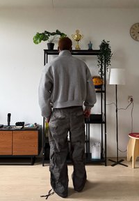 Sweat-shirt gris à col montant et coupe ample, associé à un pantalon cargo gris foncé avec plusieurs poches. Décor intérieur minimaliste.