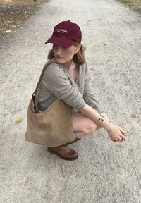 Sac hobo en daim marron avec un design incurvé et une sangle en cuir marron, porté avec un pull gris et des bijoux en bronze, sur un chemin de gravier.