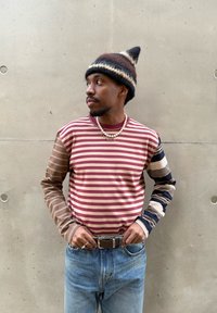 Koszula w paski z długim rękawem w odcieniach bordo, beżu i brązu; różnorodne wzory na rękawach. Noszona z wysokim stanem niebieskich dżinsów i dzianą czapką.