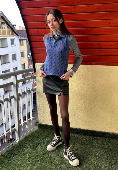 Chaleco de punto azul con patrón de trenzas sobre una camisa de rayas, combinado con una falda corta de cuero negra y zapatillas altas negras.