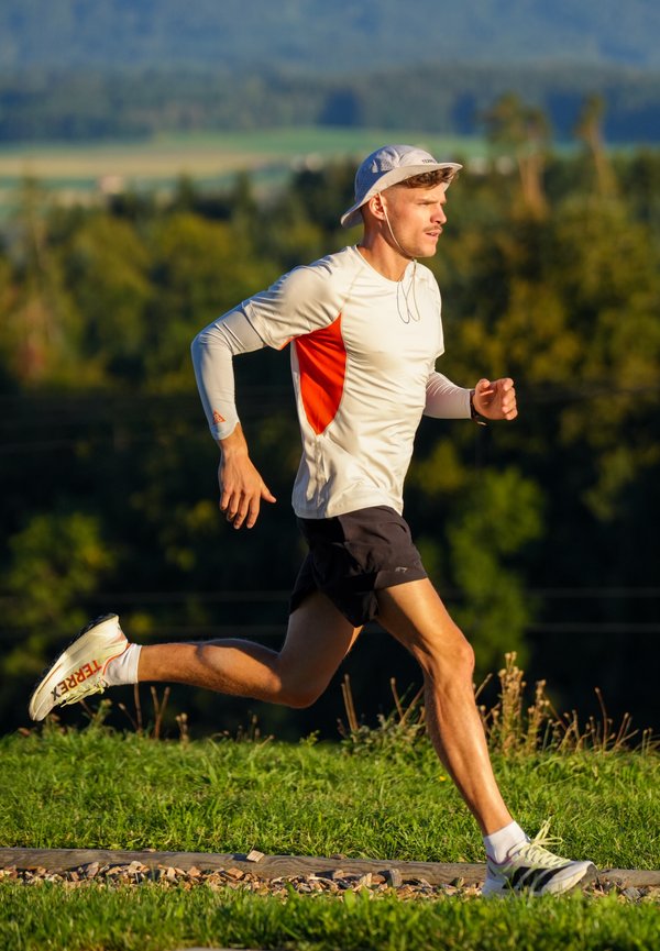 Un homme portant un chapeau de soleil, une tenue de course et des chaussures de trail court le long d’un sentier herbeux dans un parc ensoleillé.