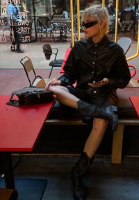 Chaqueta de camisa de cuero negro con botones, mini falda negra, botas de vaquero negras hasta la rodilla. Pequeño bolso negro sobre una mesa roja.