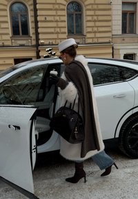 Femme en manteau de fourrure blanc et chapeau blanc tenant un café et un sac à main noir ouvre la porte d'une voiture blanche dans une rue enneigée.