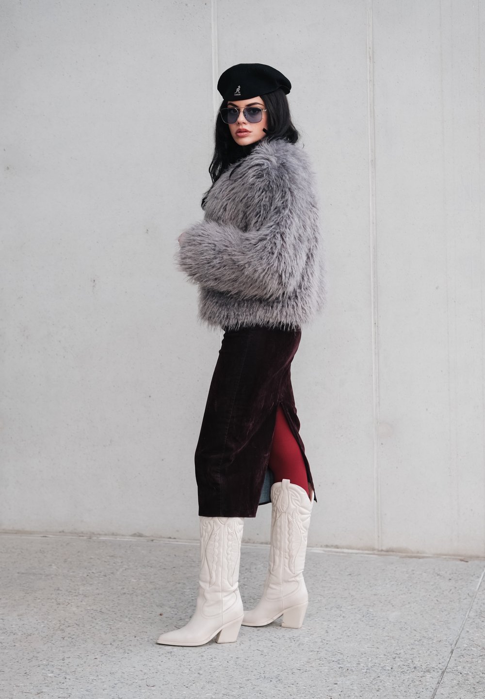 Szara kurtka z imitacji futra, bordowa aksamitna spódnica z rozcięciem, białe kowbojki, czarna beretka i okulary przeciwsłoneczne. Teksturowany i warstwowy wygląd.