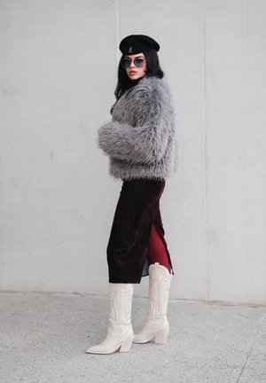 Chaqueta gris recortada de piel sintética, falda de terciopelo burdeos con abertura, botas vaqueras blancas, boina negra y gafas de sol. Look texturizado y con capas.