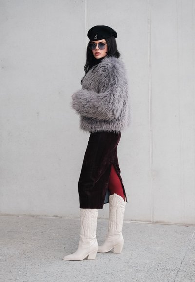 Chaqueta gris recortada de piel sintética, falda de terciopelo burdeos con abertura, botas vaqueras blancas, boina negra y gafas de sol. Look texturizado y con capas.