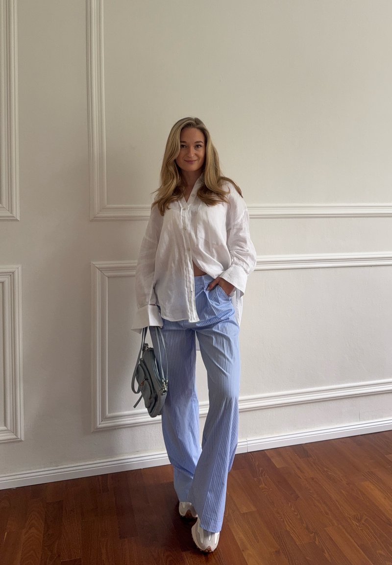 Jonge vrouw staat binnen tegen een witte geplaatste wand, draagt een wit oversized overhemd, blauwe gestreepte broek, witte schoenen en houdt een grijze handtas vast.