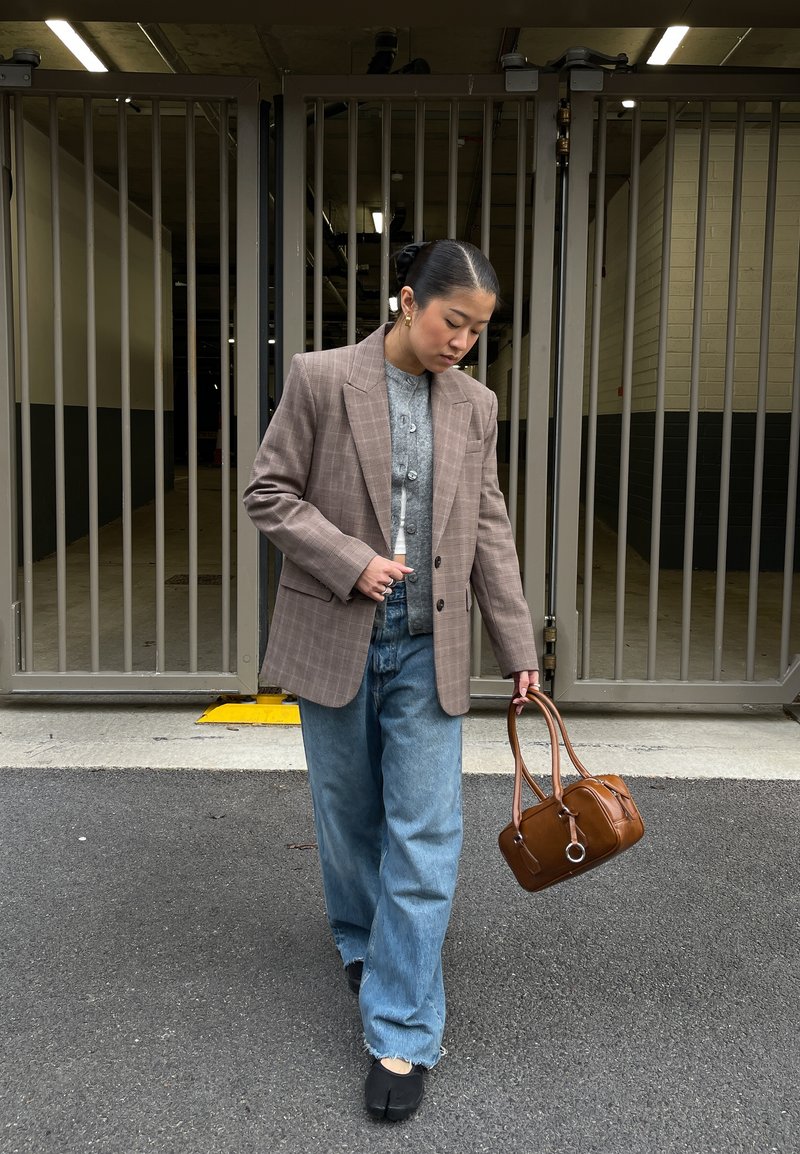 Femme portant un blazer à carreaux surdimensionné, un cardigan gris, un jean à jambes larges et des chaussures noires, tenant un sac à main en cuir marron, marchant en plein air.