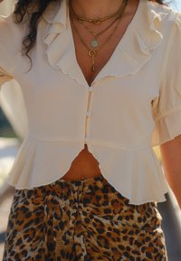 Krema-bluza s ovratnikom od nabora, kopčanjem sprijeda i kratkim rukavima; u kombinaciji s suknjom na životinjski uzorak s tamnijim smeškastim mrljama.