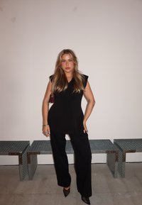 Svart skräddarsydd topp med axelvaddering, kombinerad med lösa svarta byxor och spetsiga svarta klackskor, presenterad mot en neutral bakgrund.