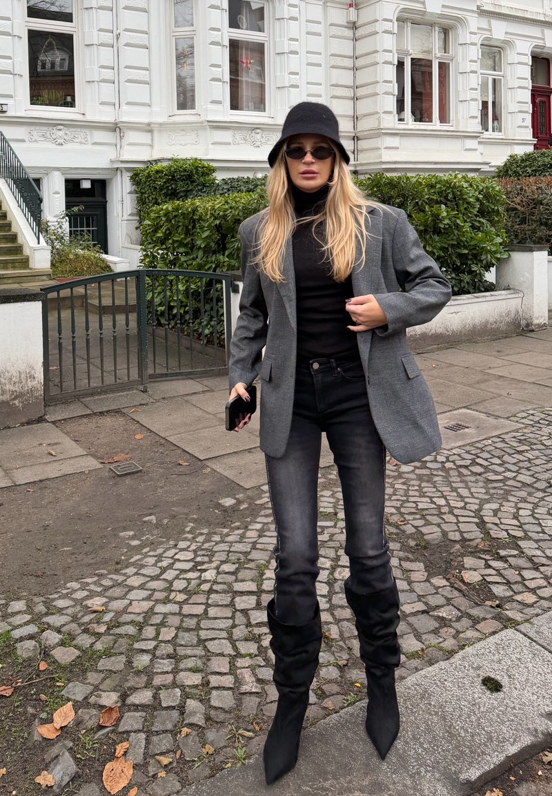 Femme portant un chapeau noir, des lunettes de soleil, un blazer gris, un col roulé noir, un jean foncé et des bottes noires, debout sur un pavé près d'un bâtiment blanc.