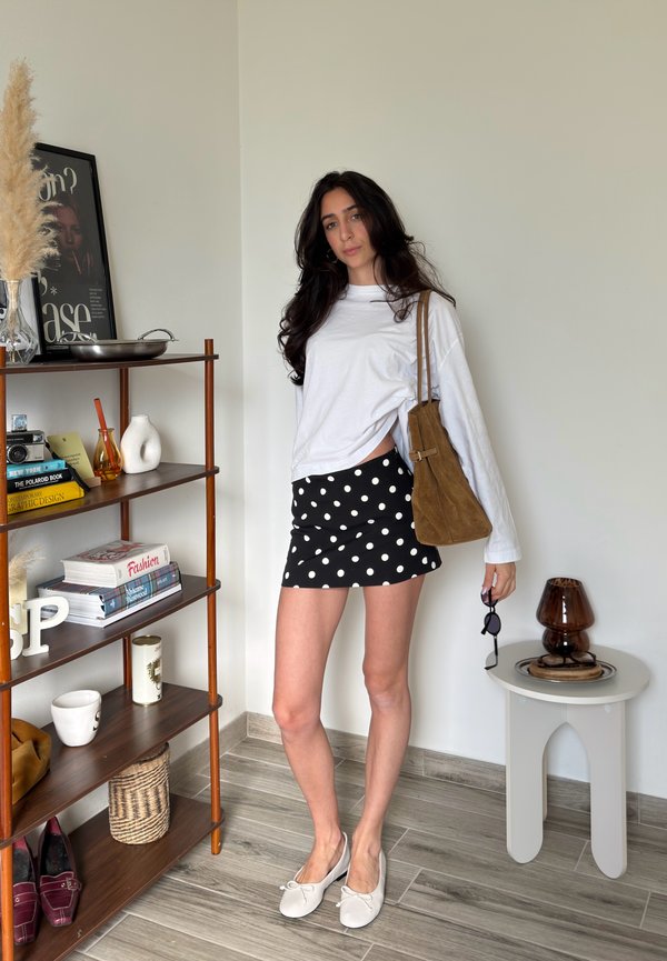Mujer con cabello largo y oscuro que lleva una camiseta blanca de manga larga, falda negra con lunares, zapatillas blancas, bolso marrón de hombro, sosteniendo gafas de sol en interiores.