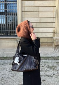 Pruun villa kapuuts, must jakk, pruun nahkviik, millel on tõmblukud, valge jänesega võtmehoidja, seismas kiviseina kõrval.