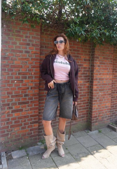 Mujer con gafas de sol, chaqueta oscura, top corto rosa, shorts de mezclilla, botas beige holgadas, sosteniendo un bolso marrón, de pie junto a una pared de ladrillos con vegetación.