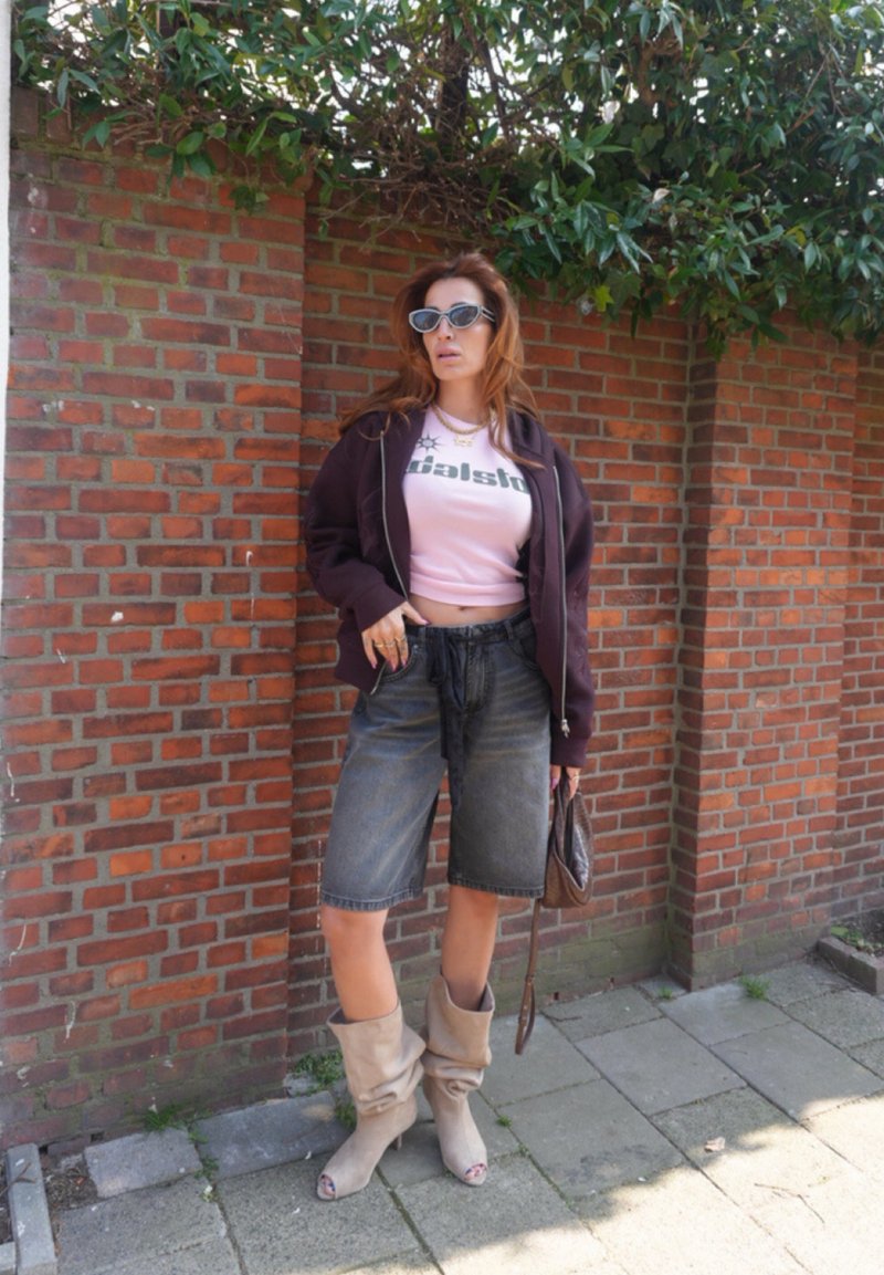 Kvinde iført solbriller, mørk jakke, pink cropped top, denimshorts, beige slouchy støvler, der holder en brun håndtaske, stående ved en murstensvæg med grønne planter.