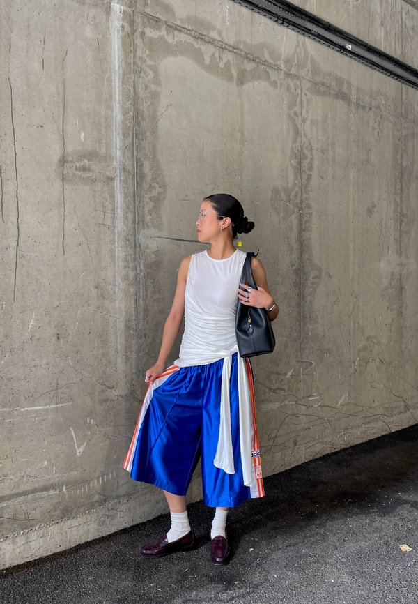 Vrouw in een witte mouwloze top, blauwe wijde broek met oranje strepen, witte sokken, donkere schoenen, draagt een zwarte tas, staat tegen een betonnen muur.