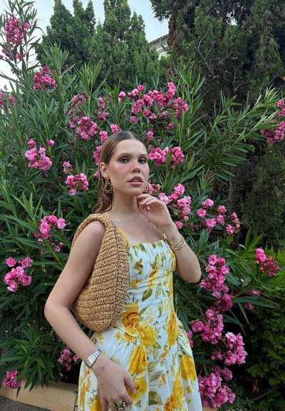Vestido amarillo con estampado floral, con un corsé ajustado y falda fluida. Bolso de mano tejido en marrón claro. Plantas con flores rosas en el fondo.