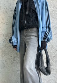 Personne portant une veste Adidas bleue avec des bandes noires, un hoodie noir à pois, un jean Adidas gris, et tenant un sac noir à pois.