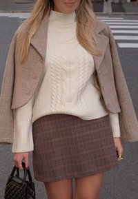 Beige houndstoothjas over een crèmekleurige kabelgebreide sweater, gecombineerd met een bruine geruite minirok; zwarte handtas met een quilted textuur.