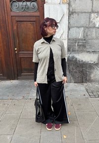 Košile s krátkými rukávy přehozená přes černý rolák, černé široké kalhoty s bílými bočními pruhy a bordó tenisky. V ruce černá taška.