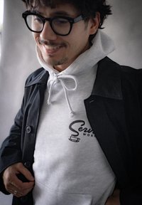 Czarny płaszcz trench na szarym hoodieu z troczkami i logo. Charakteryzuje się gładką fakturą i luźnym fasonem. Widoczne guziki na płaszczu.
