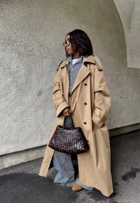 Beige trenchcoat met epauletten, zwarte knopen, gedragen over een gestreept shirt en broek. Bruine gevlochten handtas met een gestructureerde vorm.
